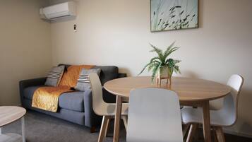 Un salon avec un canapé gris, une table ronde en bois et des chaises blanches. On peut voir une plante en pot sur la table et un tableau au mur.