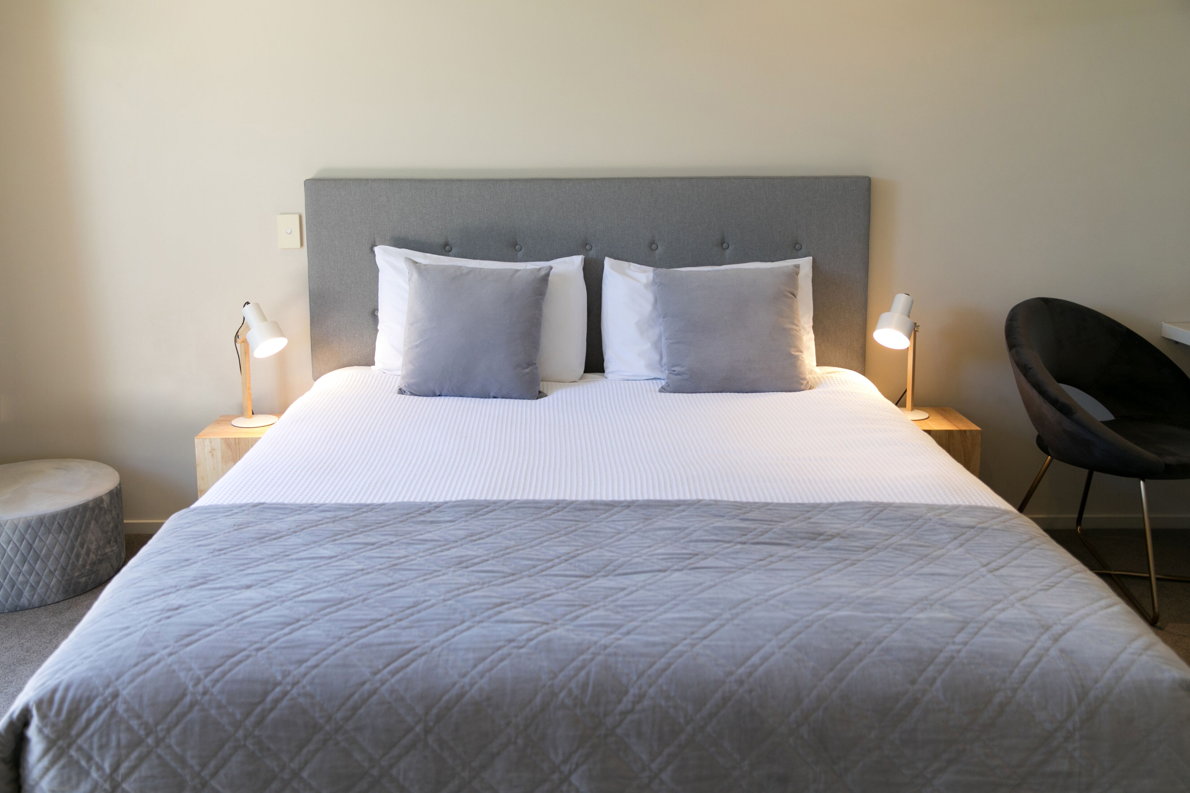 Een net opgemaakt bed met een grijs hoofdbord, twee kussens, een bedlampje, een rond voetenbankje en een moderne fauteuil.