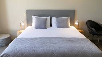 Een net opgemaakt bed met een grijs hoofdbord, twee kussens, een bedlampje, een rond voetenbankje en een moderne fauteuil.