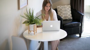 A person sitting at a round white table with a laptop, a potted plant, and a black leather couch in the background. - Alpine Lake Motor Lodge (Taupo)