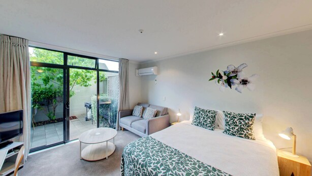 A modern hotel room with a large bed, a sofa, a TV, and a balcony with a view of greenery. - Alpine Lake Motor Lodge (Taupo)