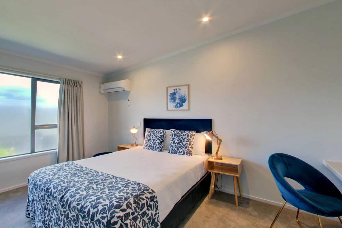 Una habitación de hotel con una cama grande, un escritorio, una silla, una mesita de noche con una lámpara, un equipo de aire acondicionado y una ventana con vistas.