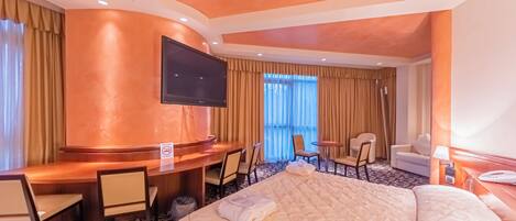 A hotel room with a large bed, a desk with chairs, a television, and a window with curtains.
