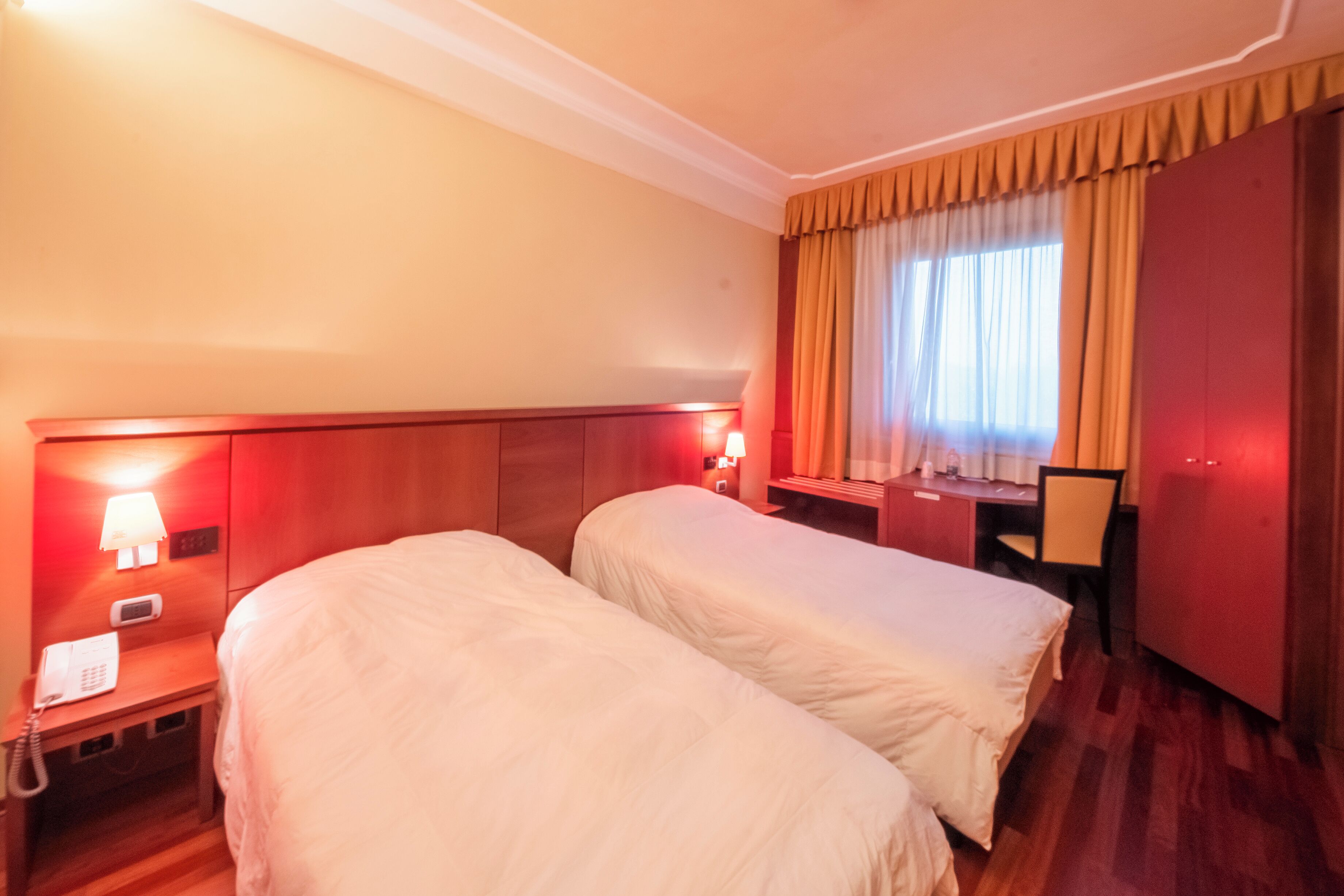 A hotel room with two single beds, a desk, a chair, a telephone, and a window with curtains.