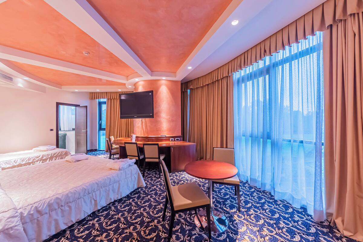 Ein Hotelzimmer mit zwei Betten, einem Schreibtisch, einem Stuhl, einem Fernseher und großen Fenstern mit Vorhängen.