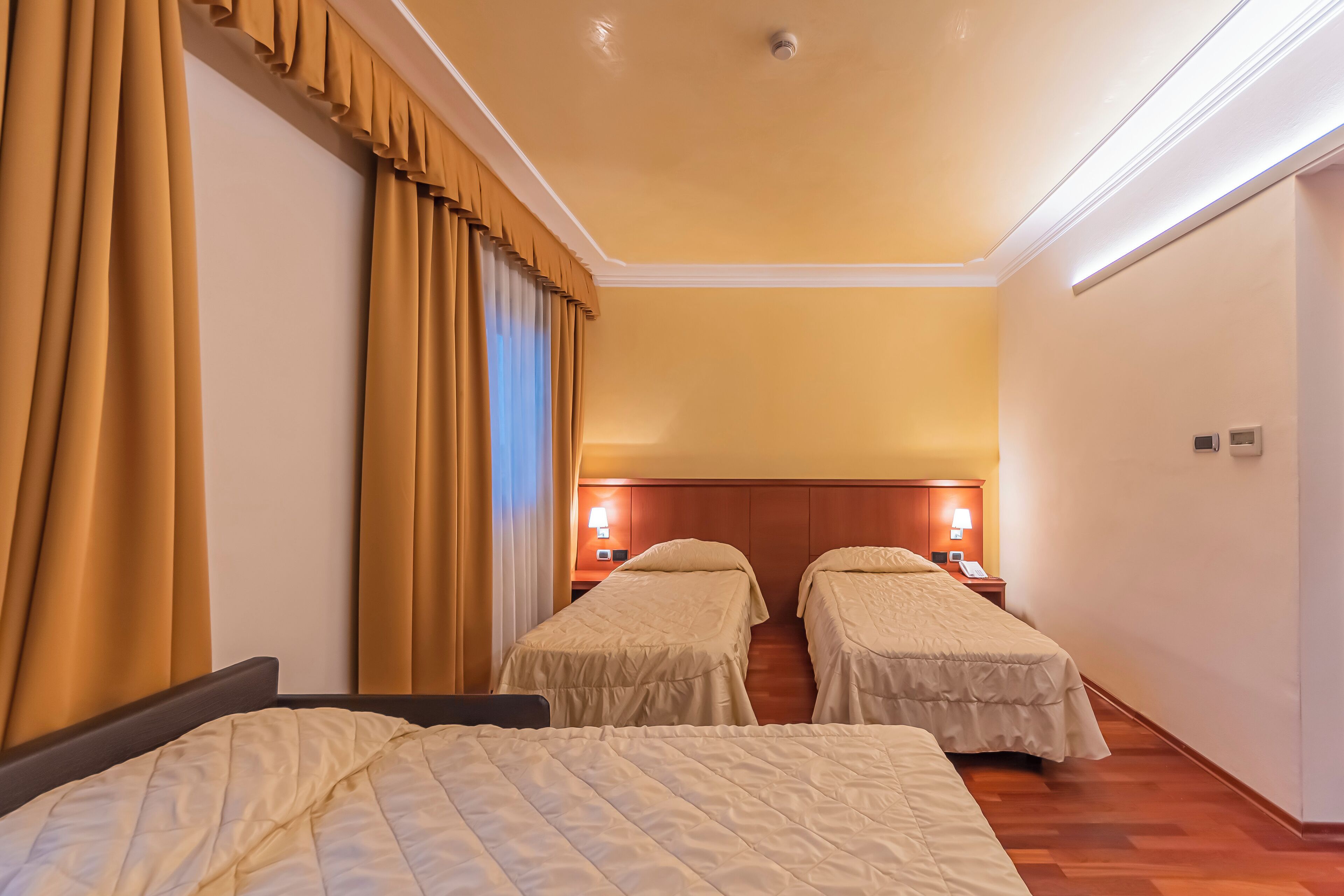 Una camera d'albergo con due letti singoli, un letto grande, una testiera in legno, un comodino con una lampada e una finestra con tende.