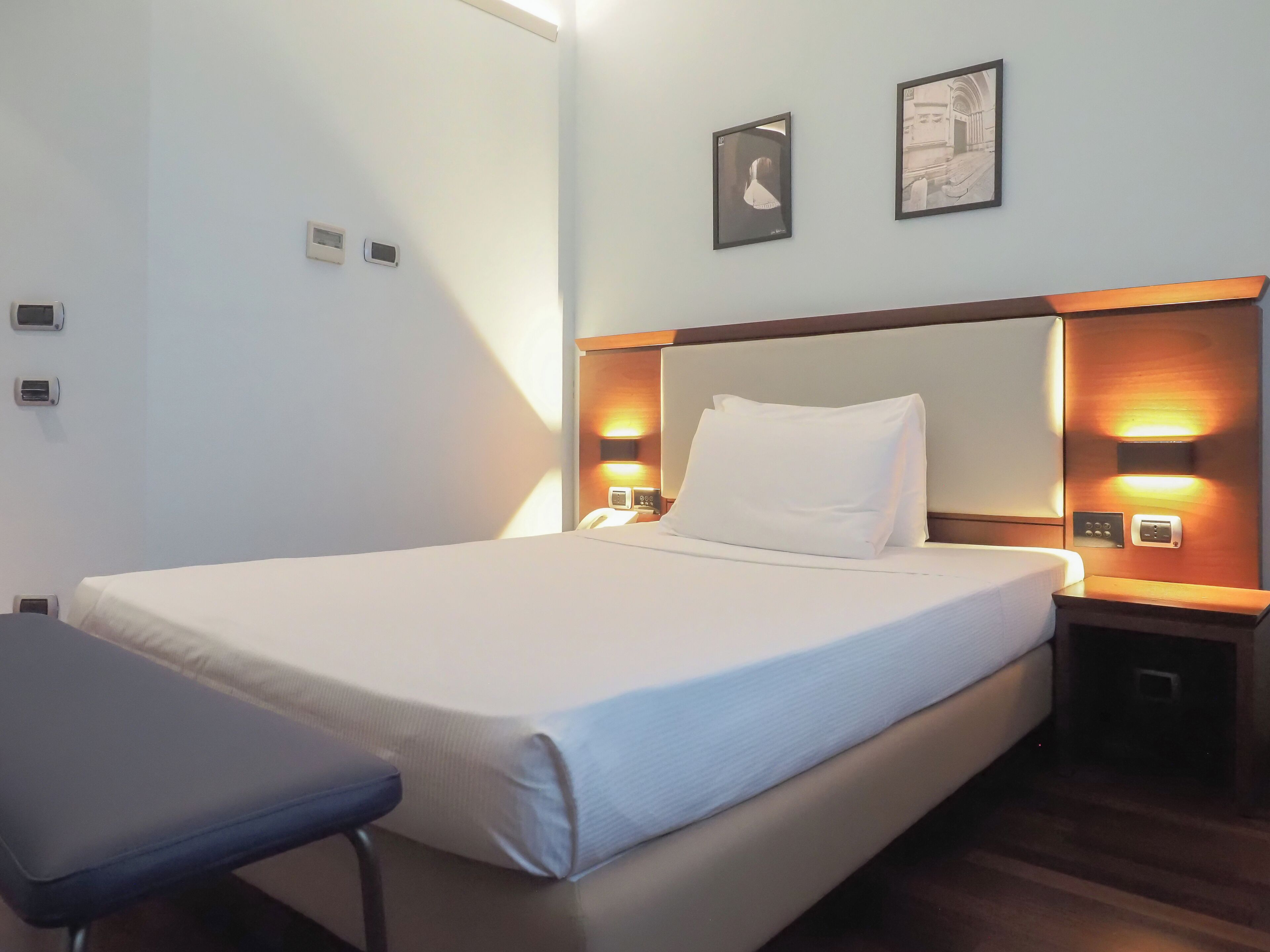 Una cama bien tendida con sábanas blancas, cabecera de madera, mesitas de noche con lámparas y dos cuadros en la pared.