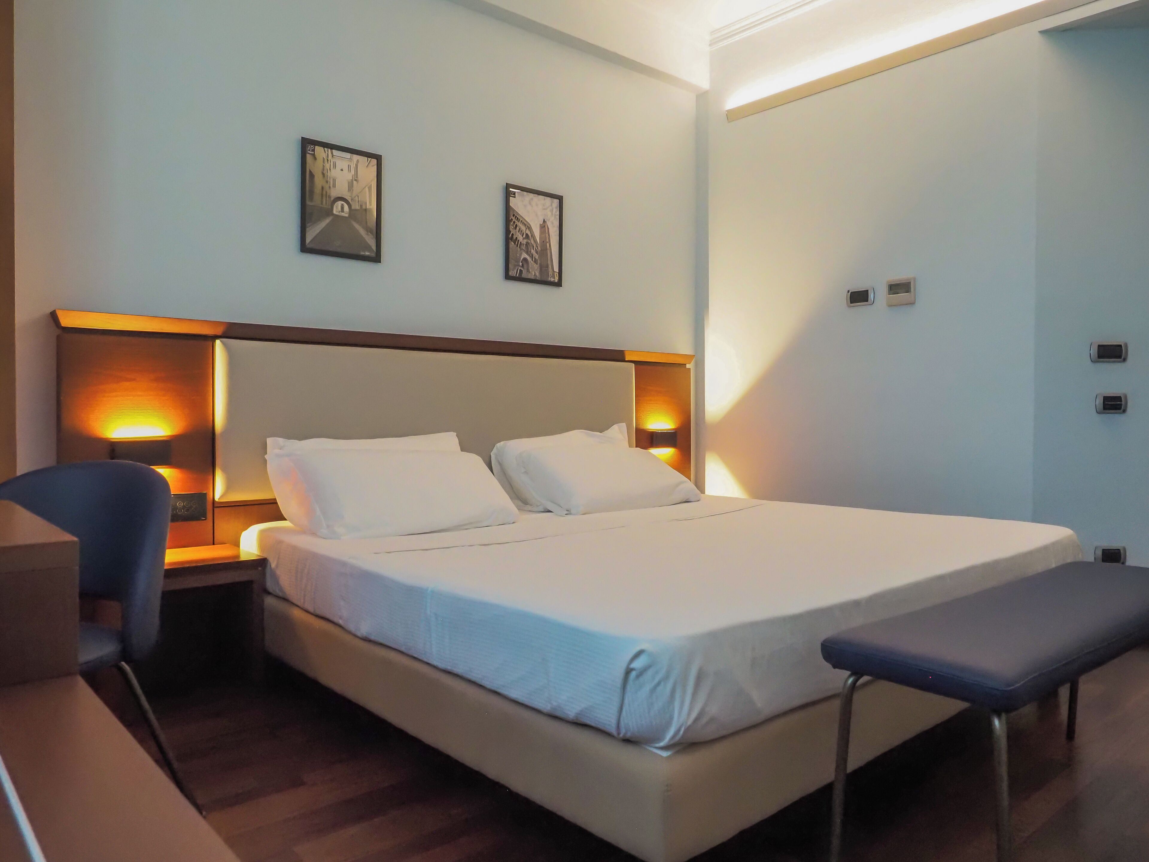 Een hotelkamer met een bed, nachtkastjes, een stoel en een bankje. Twee ingelijste foto's hangen aan de muur.