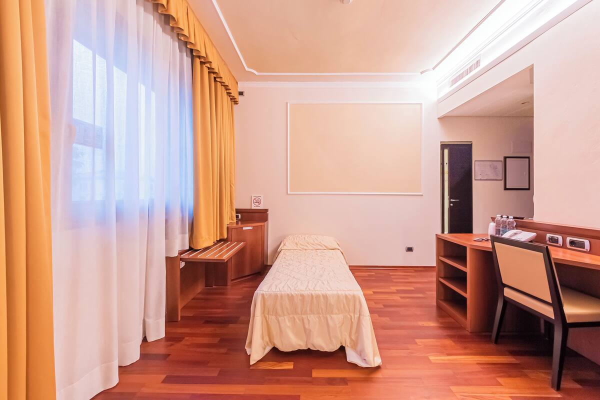 Ein Hotelzimmer mit einem Bett, einem Schreibtisch und einem Sessel. Es gibt ein Fenster mit Vorhängen, eine Tür und ein an der Wand befestigtes Regal mit Telefon und einem kleinen Tisch.