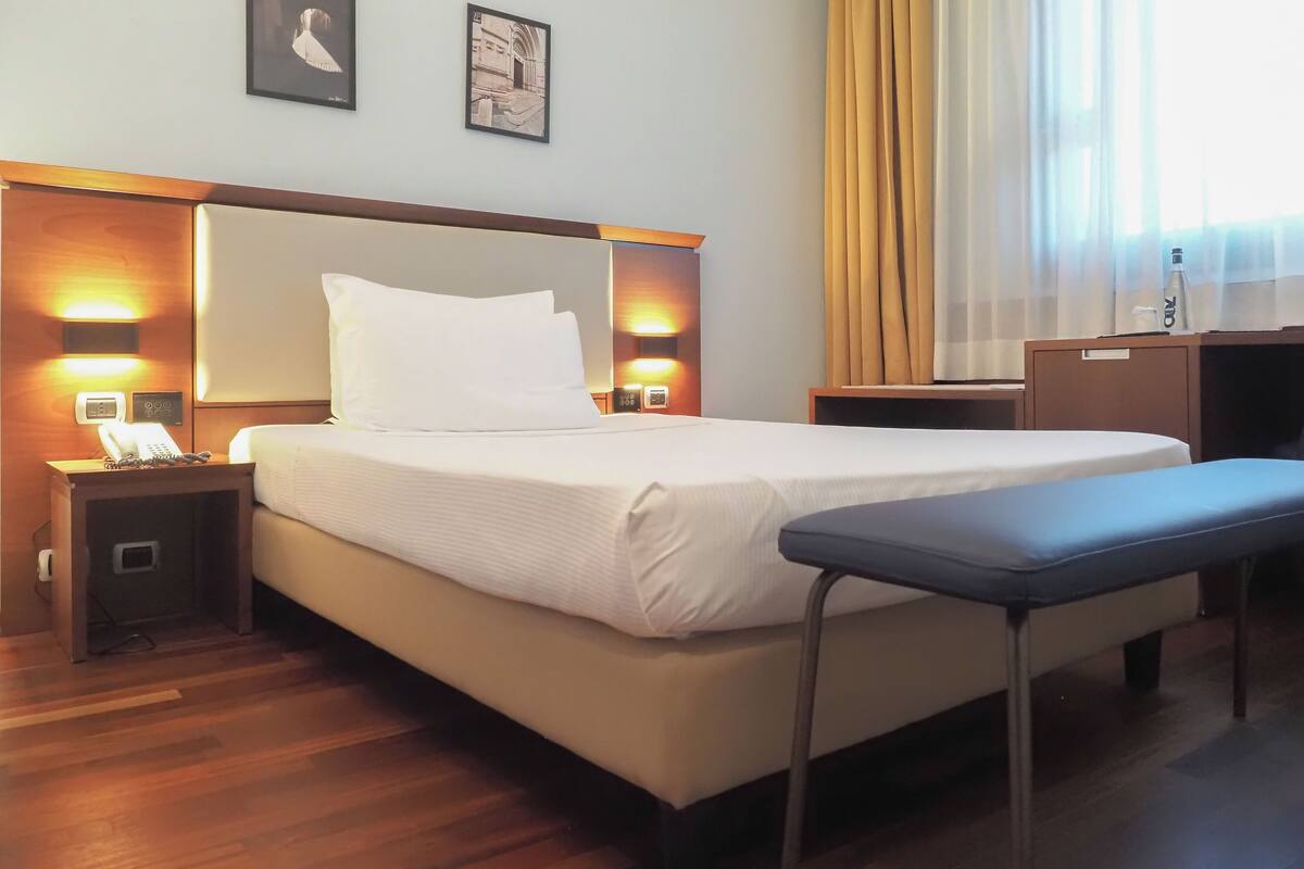 Ein Hotelzimmer mit Bett, Nachttischen, Schreibtisch und Stuhl. An der Wand hängen zwei gerahmte Bilder, auf dem Nachttisch ein Telefon und auf dem Schreibtisch eine Flasche.