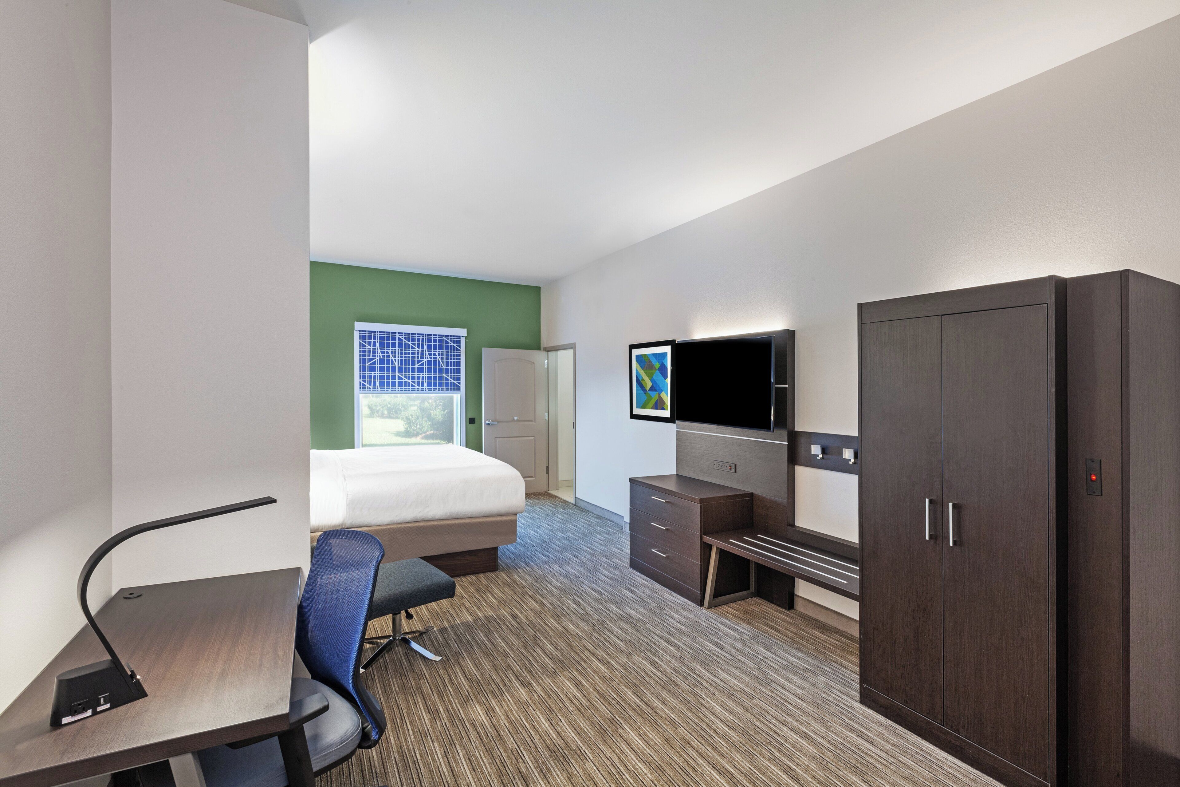 Habitación de hotel con cama, escritorio, silla, televisor, armario y ventana con persianas.