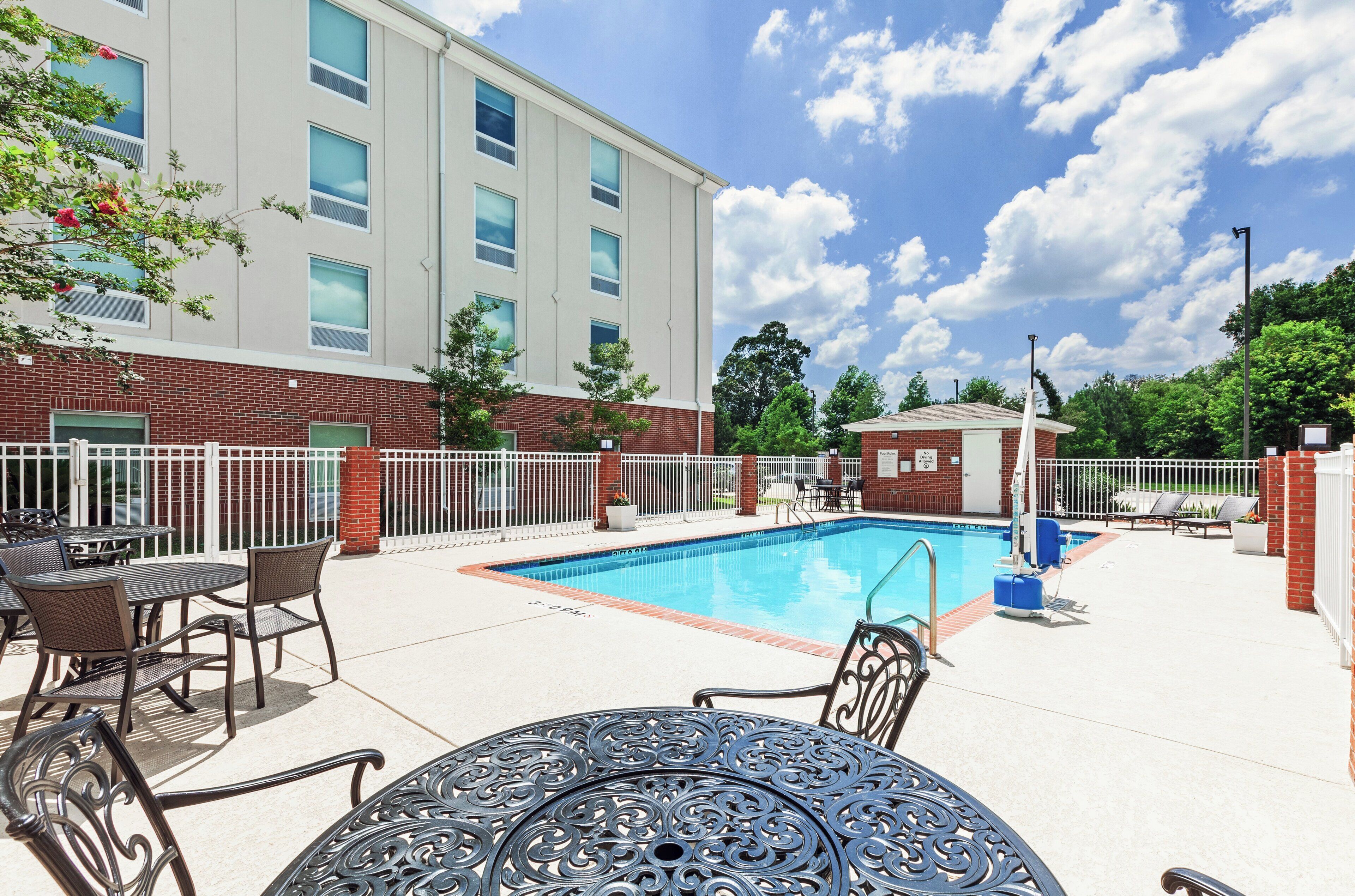 A swimming pool with a white fence, outdoor seating, and a building with multiple windows in the background.
