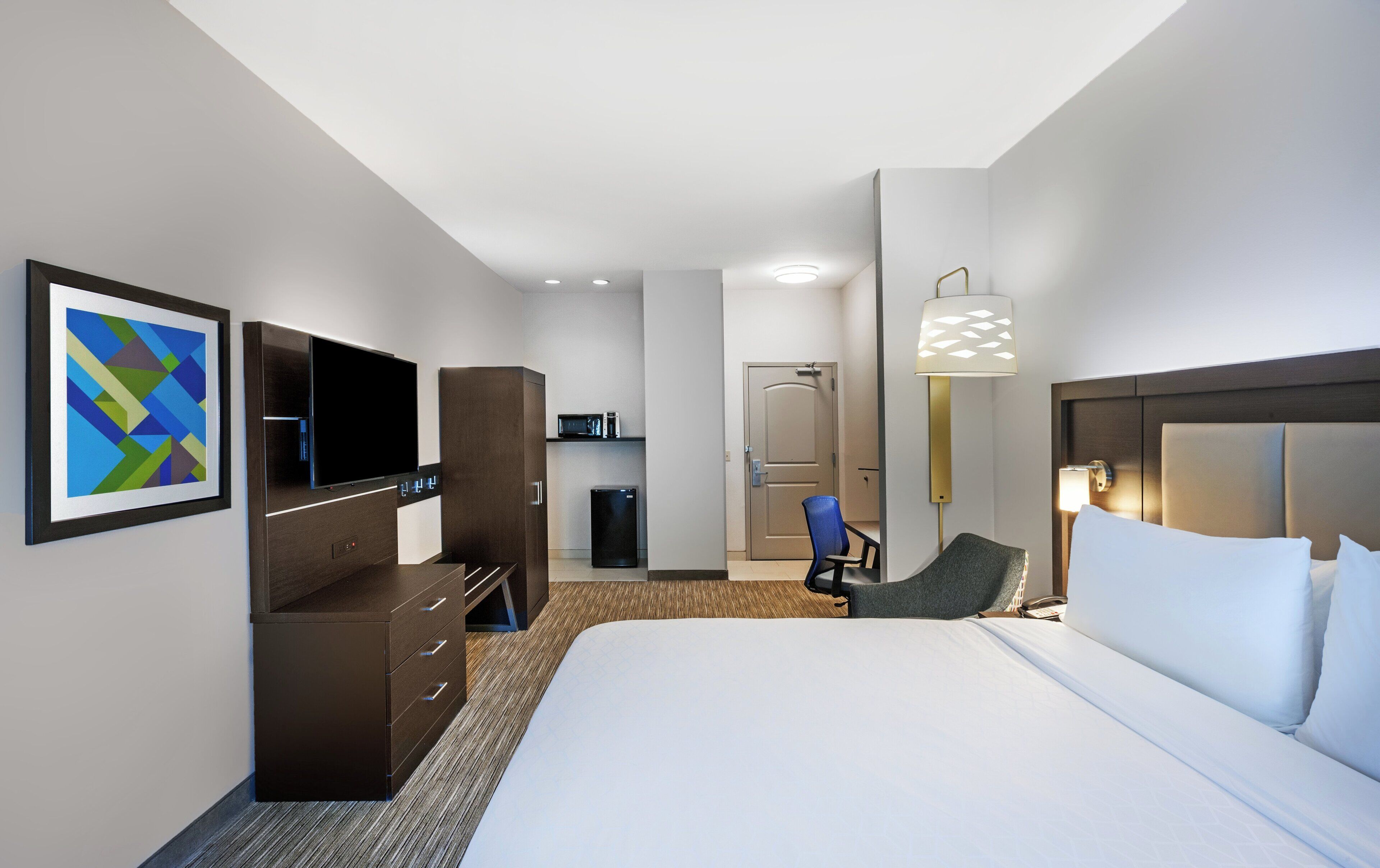 Ett hotellrum med en säng, ett skrivbord, en stol, en tv, en minibar och en dörr.