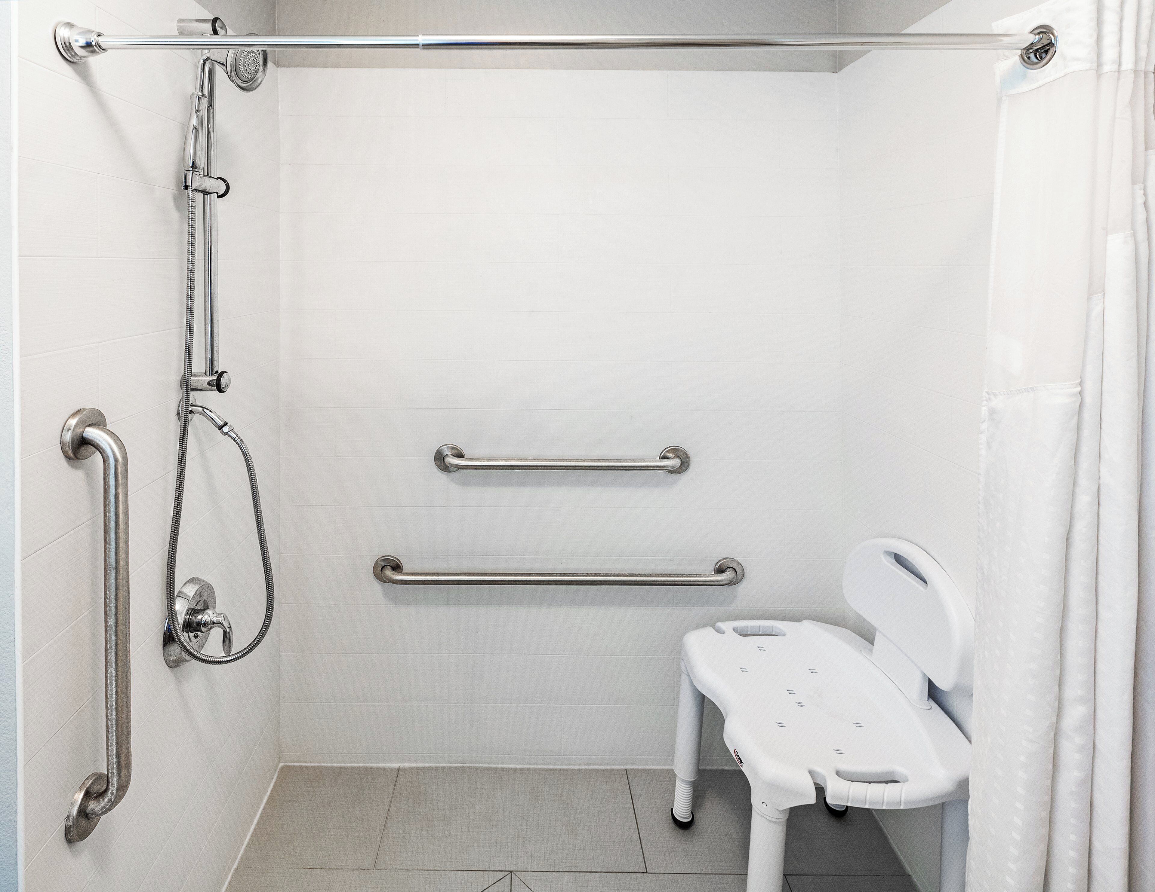 A bathroom with white tiled walls, a shower with a handheld showerhead, grab bars, and a white toilet with a safety seat.