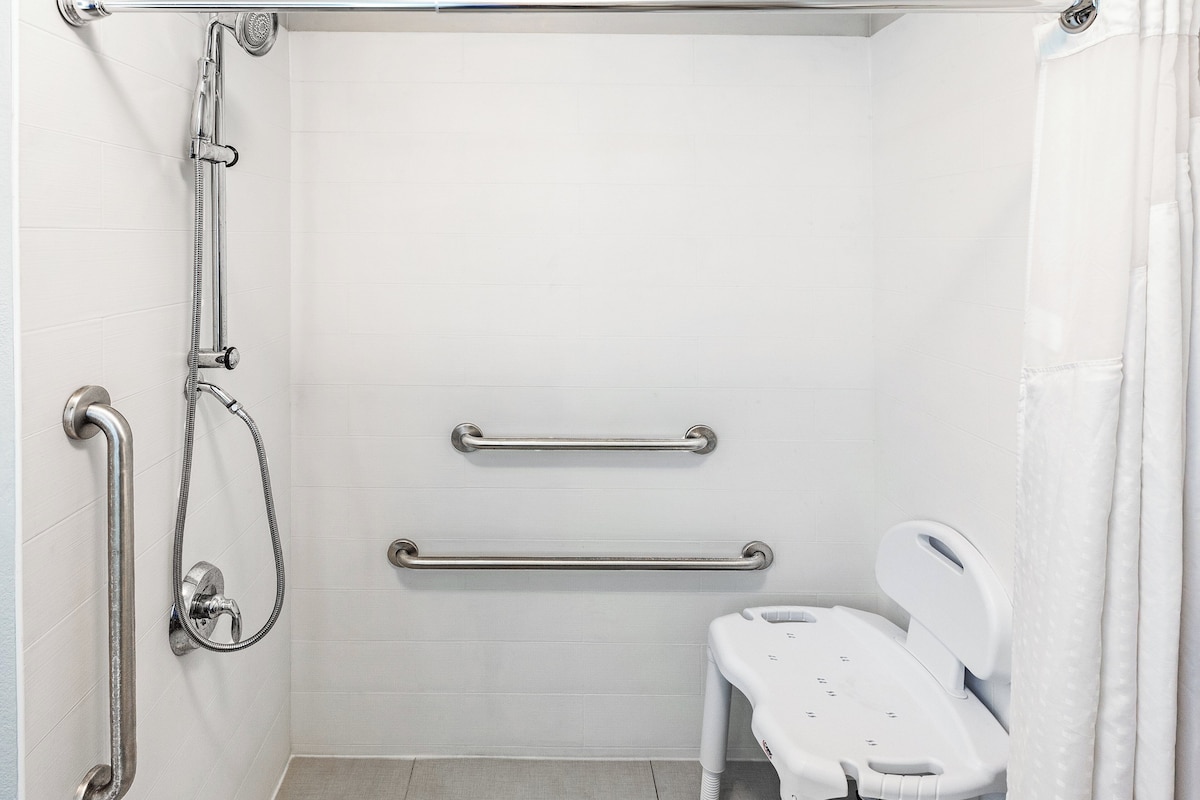 A bathroom with white tiles, a shower with grab bars, a white toilet, and a shower seat.
