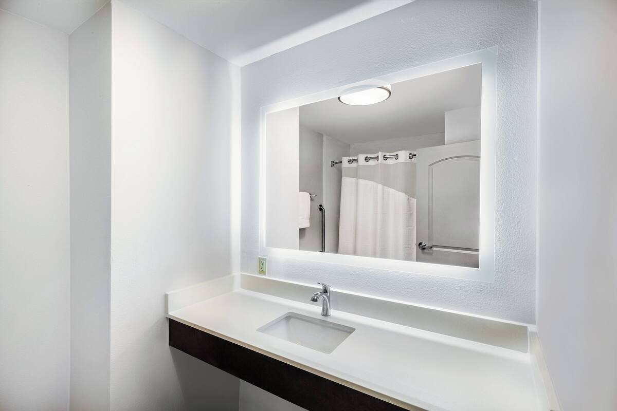 A modern bathroom with a white vanity, a mirror, and a shower visible in the reflection.