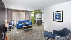 Quarto de hotel com um sofá azul, uma escrivaninha com cadeira, uma mesa pequena e uma poltrona estampada.