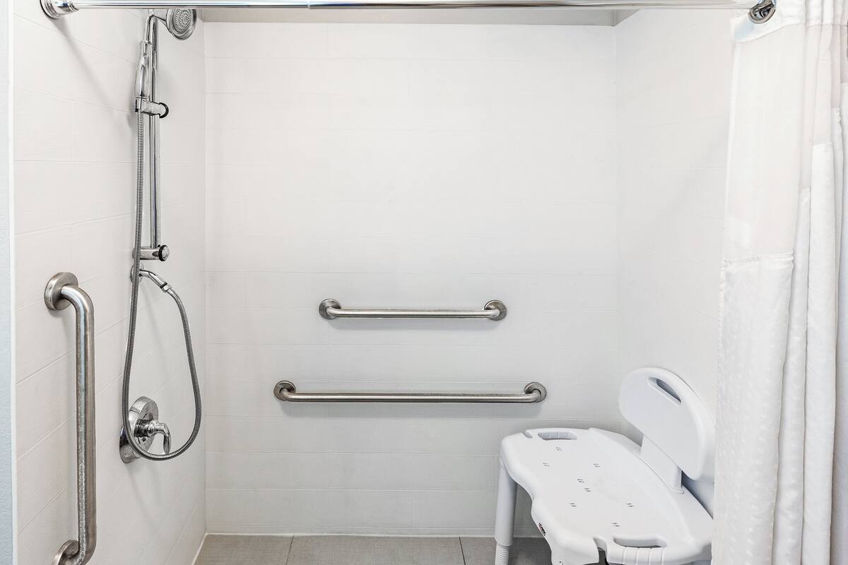 A bathroom with white tiles, a shower with grab bars, a white toilet, and a shower seat.