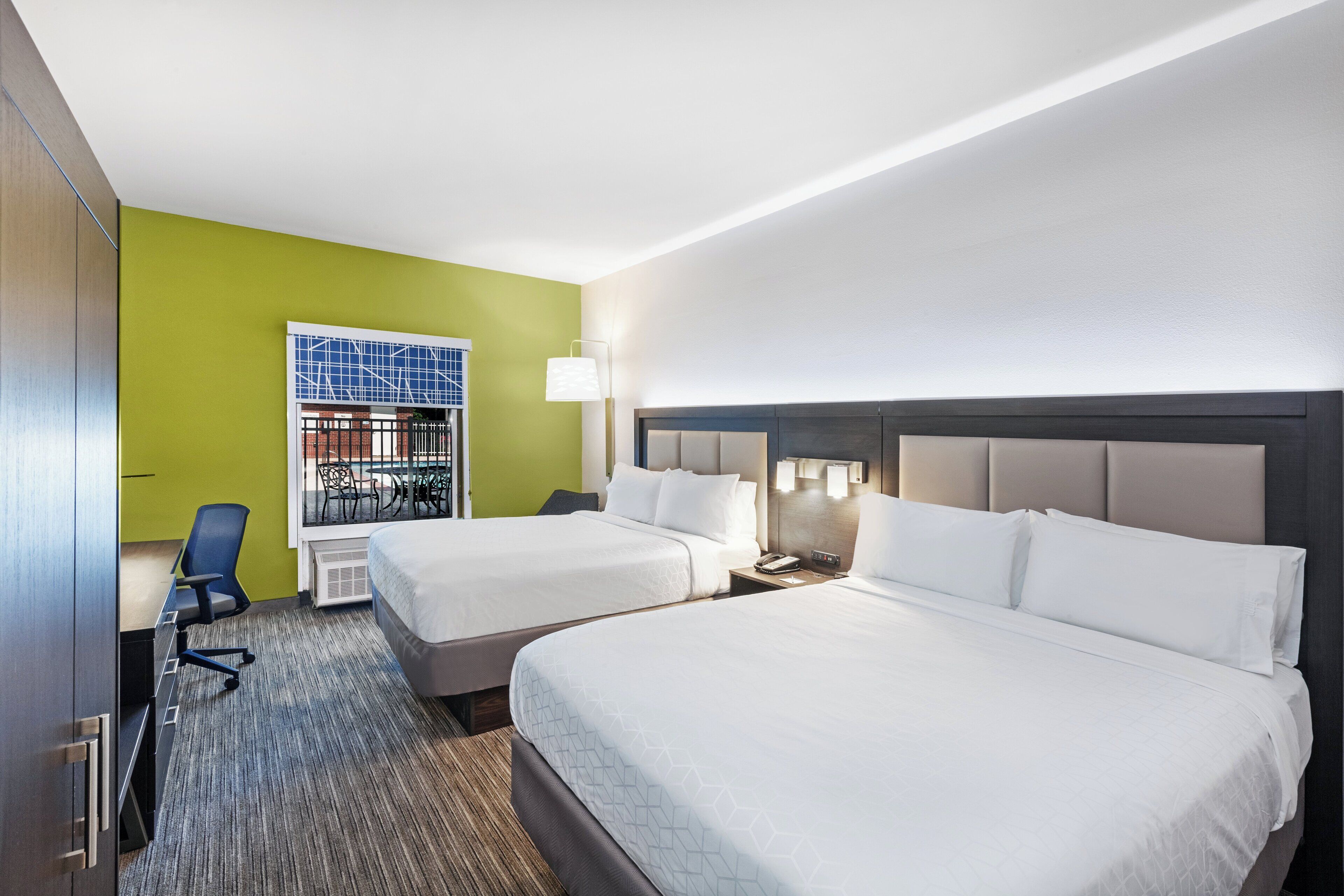 Ein Hotelzimmer mit zwei Betten, einem Schreibtisch, einem Stuhl und einem Fenster mit Blick auf ein Gebäude.