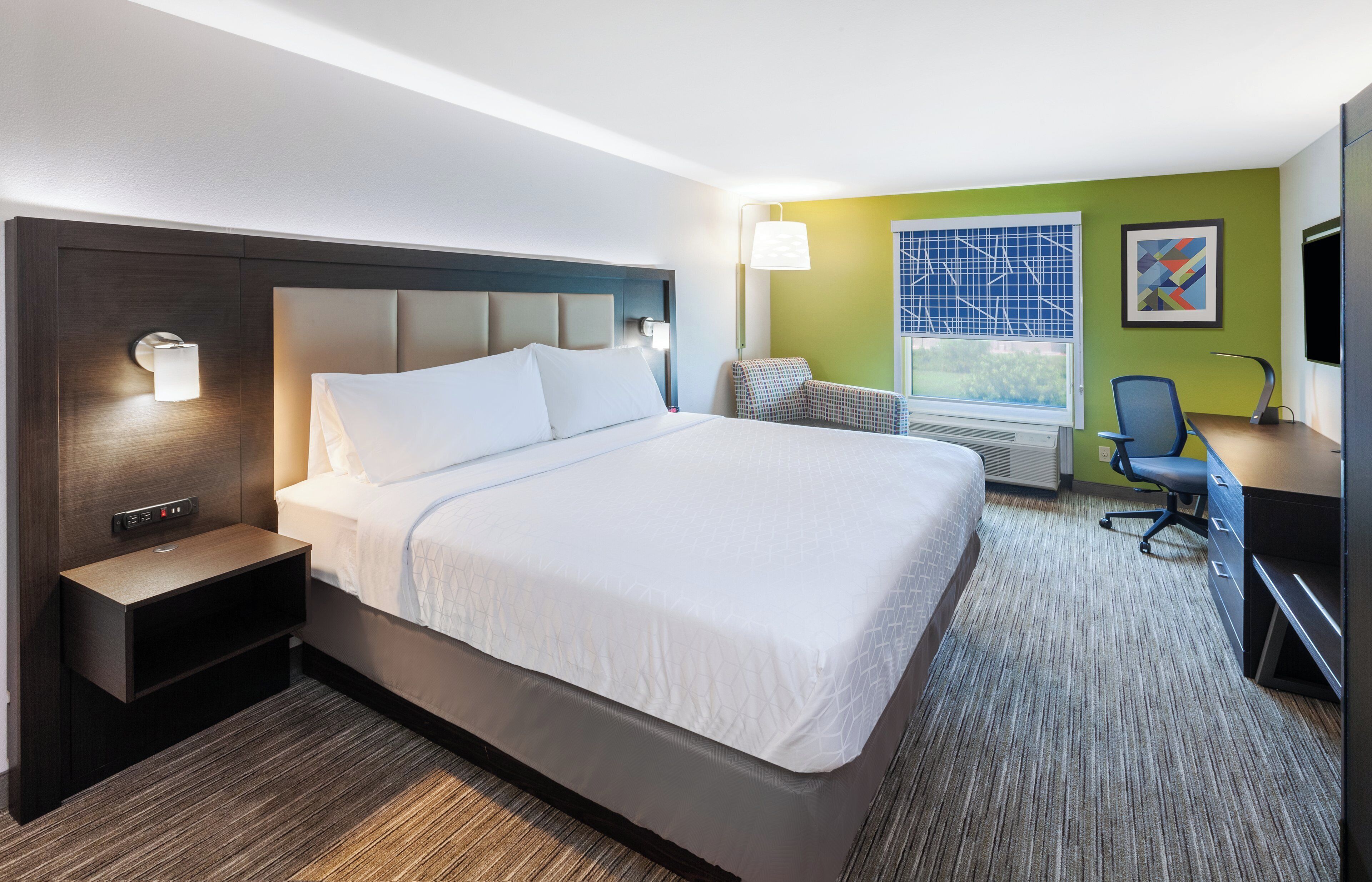 Une chambre d’hôtel avec un grand lit, des tables de chevet, un bureau avec une chaise, une télévision et une fenêtre avec des stores.