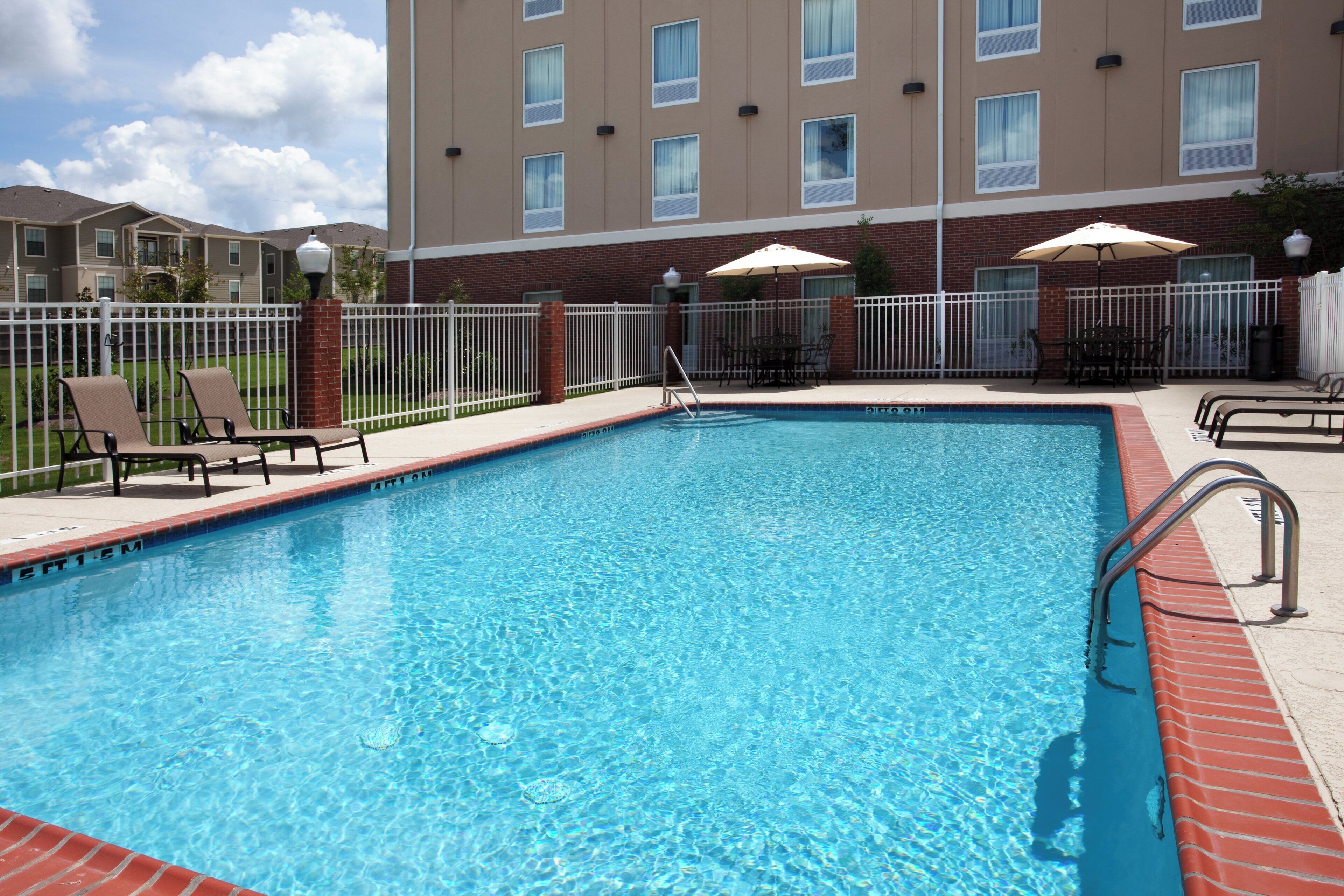 A rectangular swimming pool with a metal ladder on one side, surrounded by a white fence. There are lounge chairs and a table with an umbrella on the poolside.