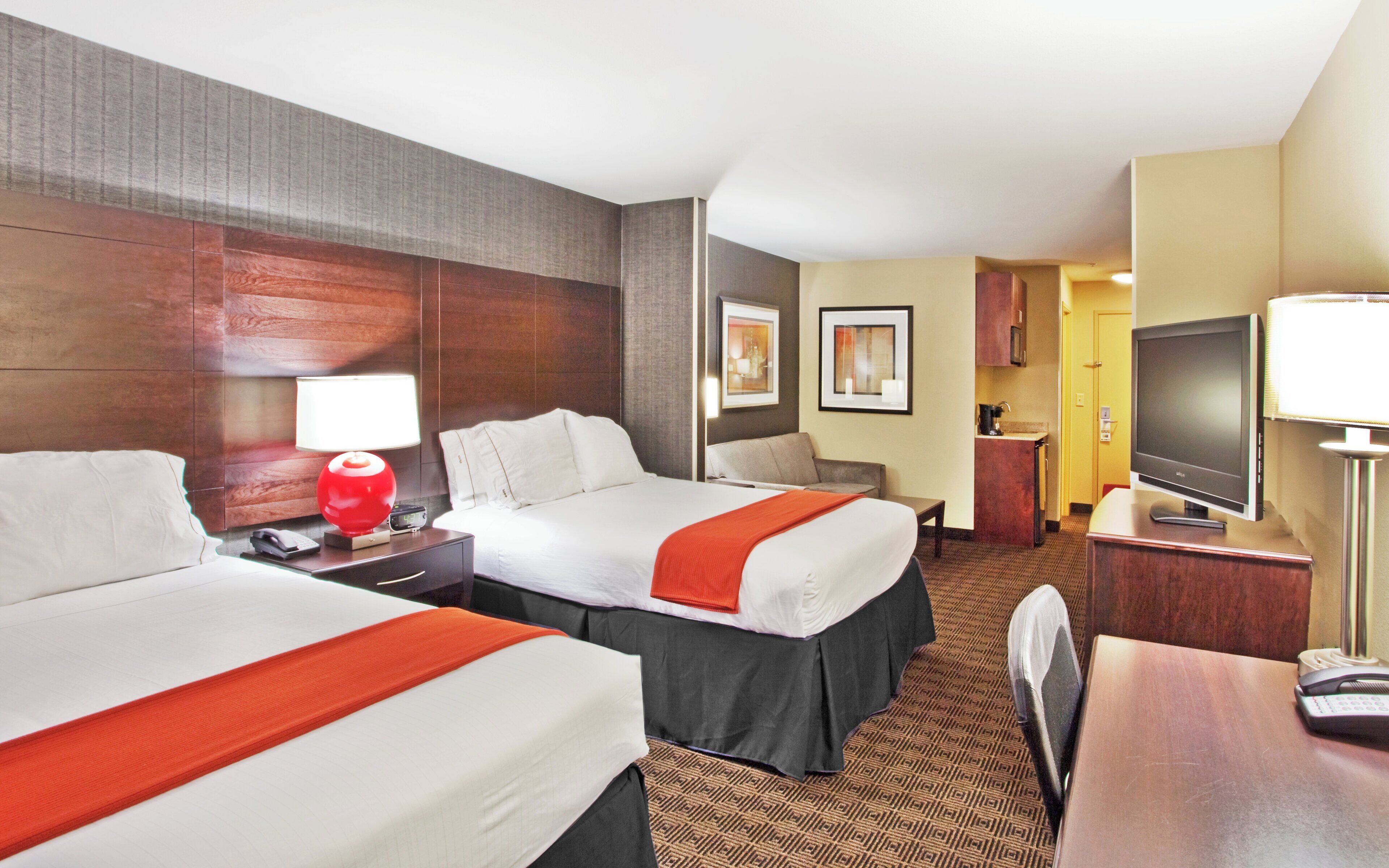 A hotel room with two beds, a desk, a television, and a small kitchenette.