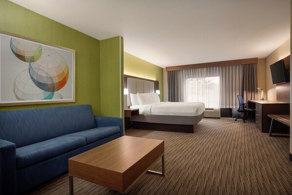 A hotel room with a bed, a blue sofa, a wooden coffee table, a desk with a chair, and a television on the wall. - Holiday Inn Express Hotel & Suites Atlanta-Cumming by IHG (Cumming)