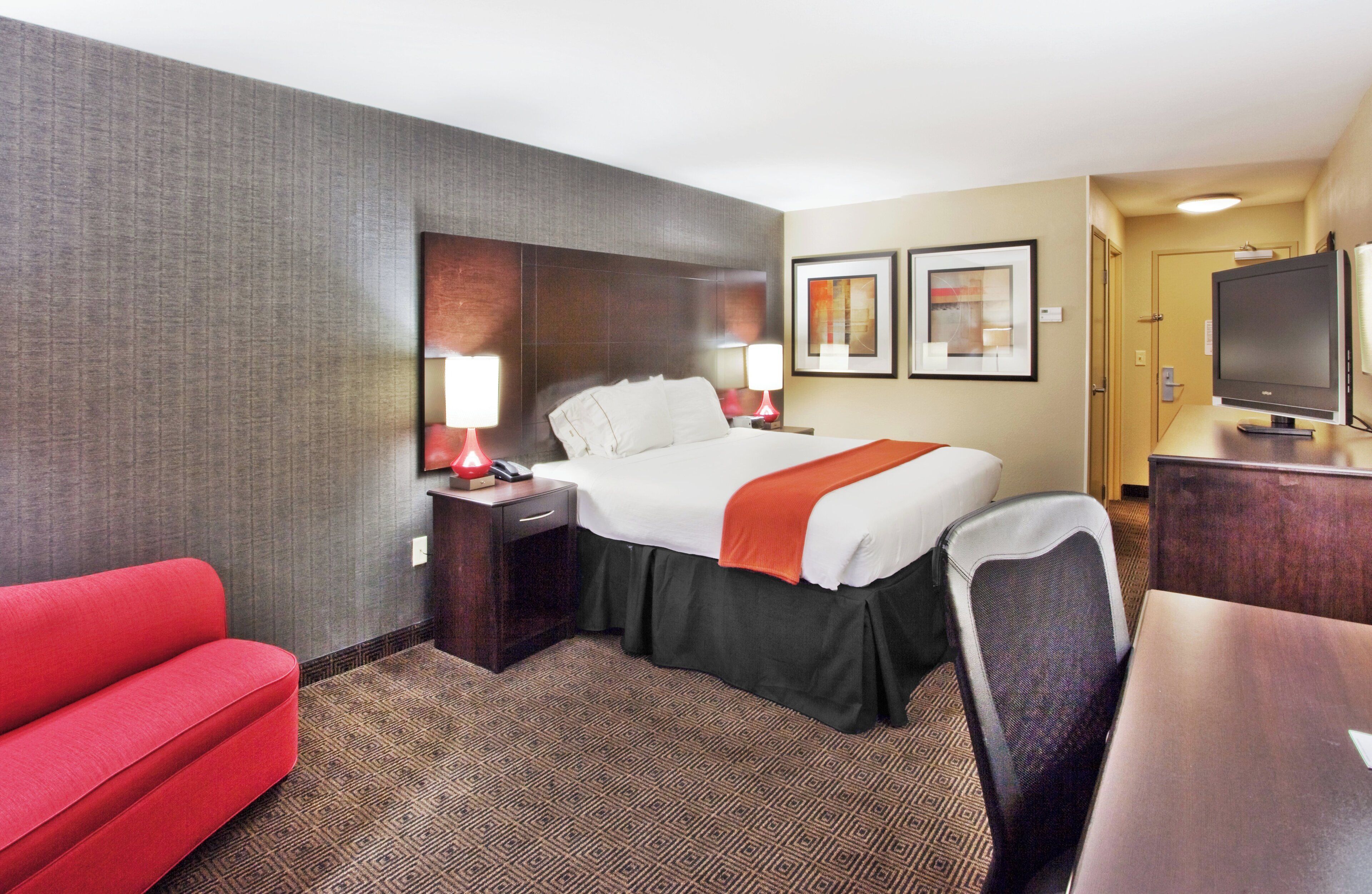 A hotel room with a bed, a desk, a TV, a red sofa, and two framed pictures on the wall.
