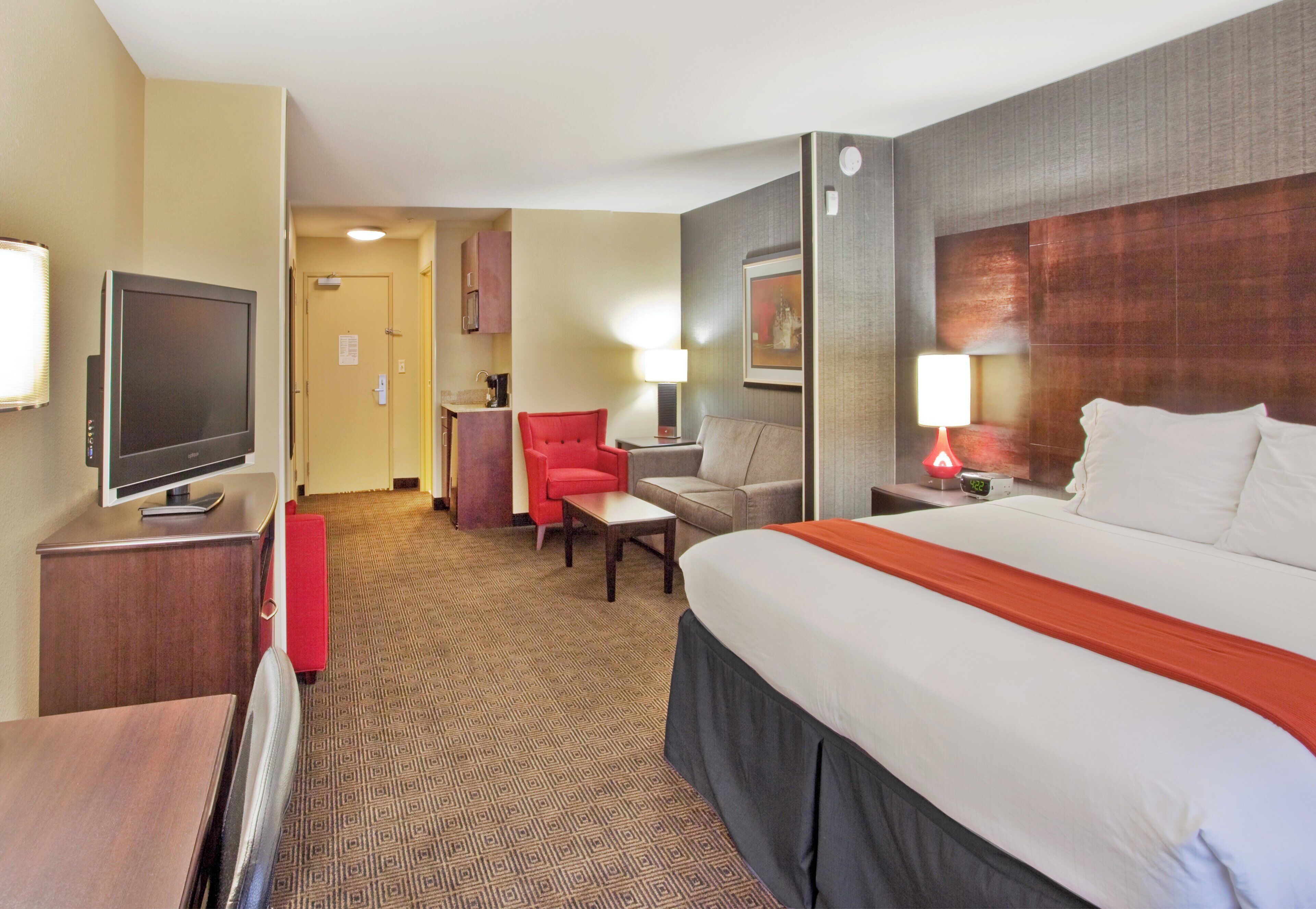 A hotel room with a large bed, a television, a red chair, a sofa, and a desk.