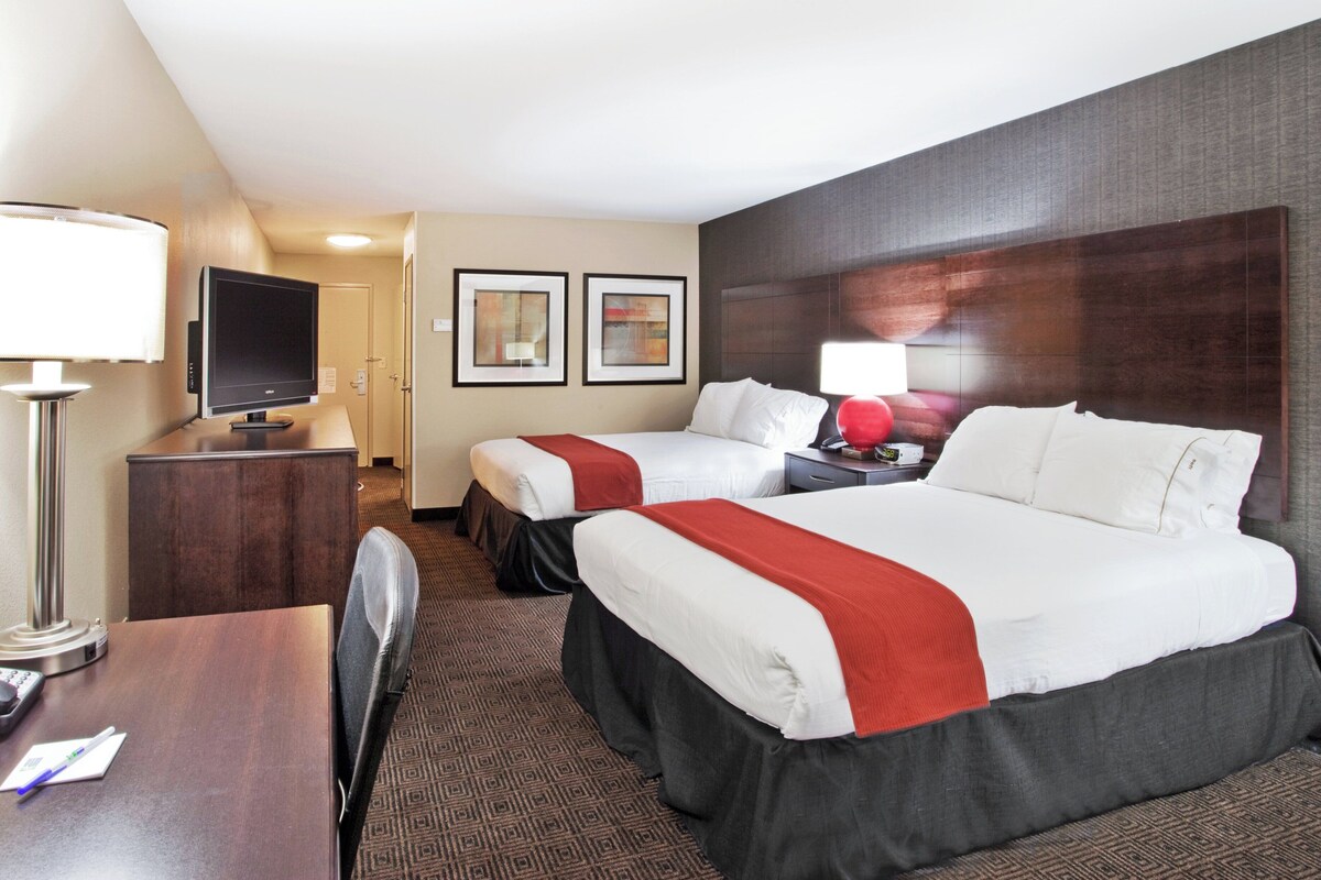 A hotel room with two beds, a desk with a lamp, a TV, and framed artwork on the wall.