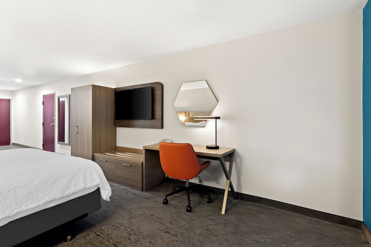 A hotel room with a bed, a desk, an orange office chair, a flat-screen TV, and a lamp.