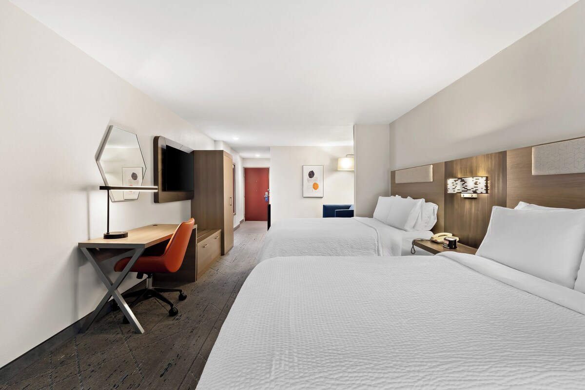 Una habitación de hotel con escritorio, dos camas, un espejo y televisor montado en la pared.