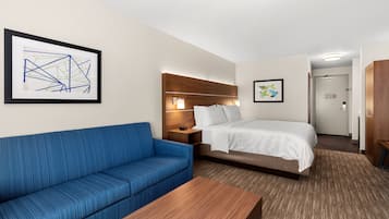 Habitación de hotel con sofá azul, cama con ropa de cama blanca, mesita de noche de madera y un cuadro abstracto enmarcado en la pared.