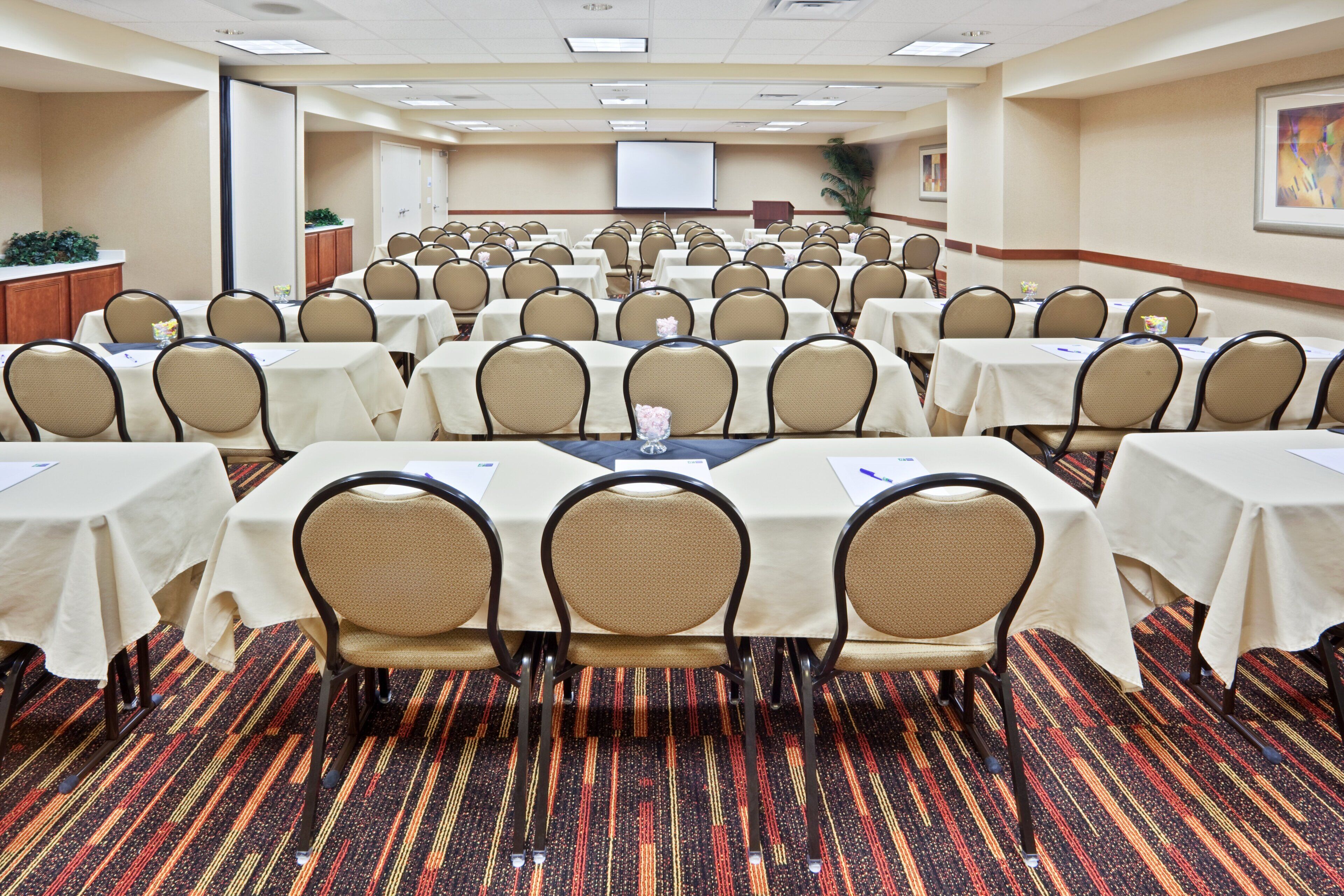 Une salle de conférence équipée de plusieurs tables et chaises, d’un écran de projection et d’un sol recouvert de moquette.