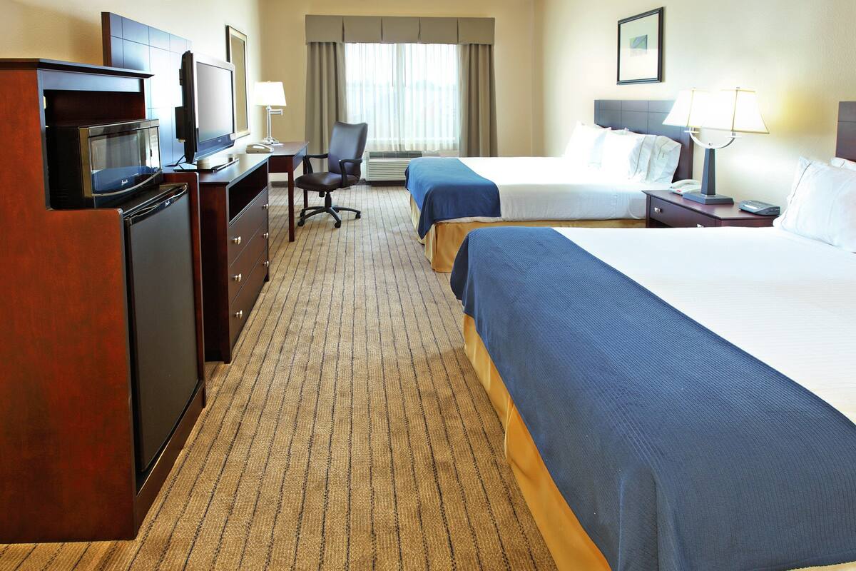 A hotel room with a desk, chair, television, minibar, two beds, and a window with curtains.