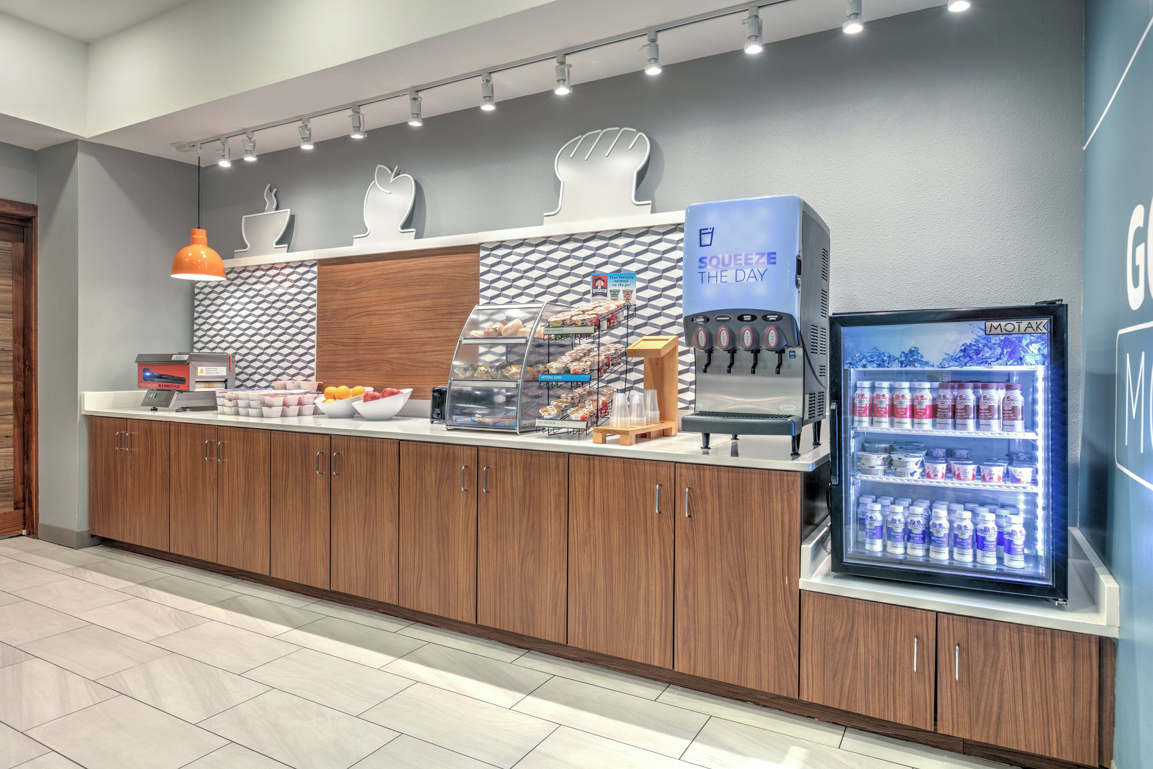 Un área de desayuno autoservicio con una máquina de café, dispensador de bebidas refrigeradas y una vitrina con pasteles.