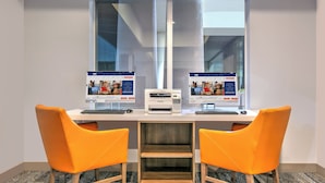 Un espace de bureau moderne comprenant deux bureaux, chacun équipé d’un écran d’ordinateur, d’un clavier et d’une imprimante. On y trouve deux chaises orange et une petite étagère en bois placée entre les bureaux.