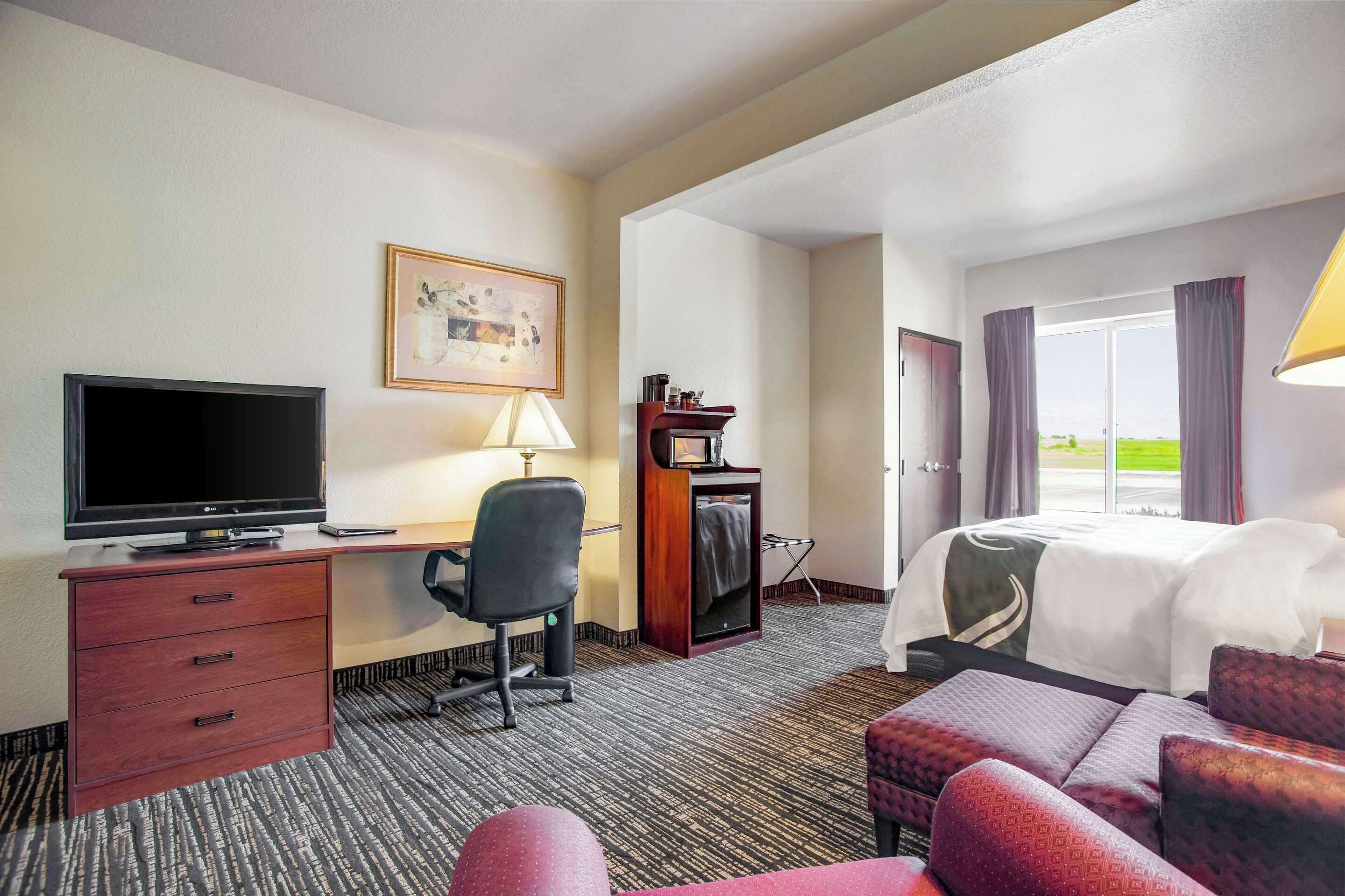 A hotel room with a bed, a desk with a TV, a chair, a lamp, and a window with curtains.
