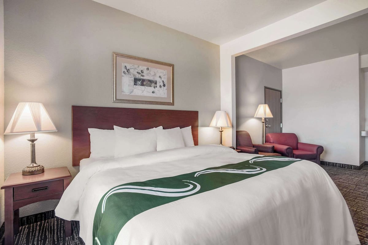 Une chambre d’hôtel avec un grand lit, deux tables de chevet avec des lampes, un tableau encadré au mur et un fauteuil rouge dans un coin.