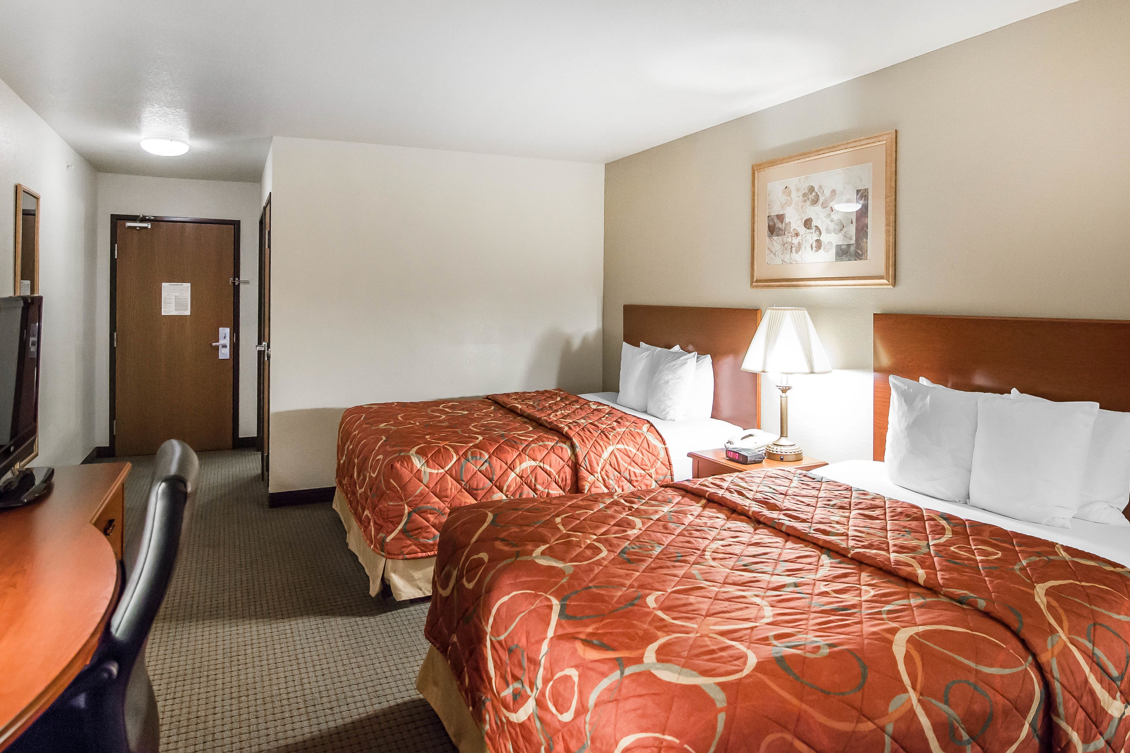 A hotel room with two beds, a desk with a chair, a lamp, and a framed picture on the wall.