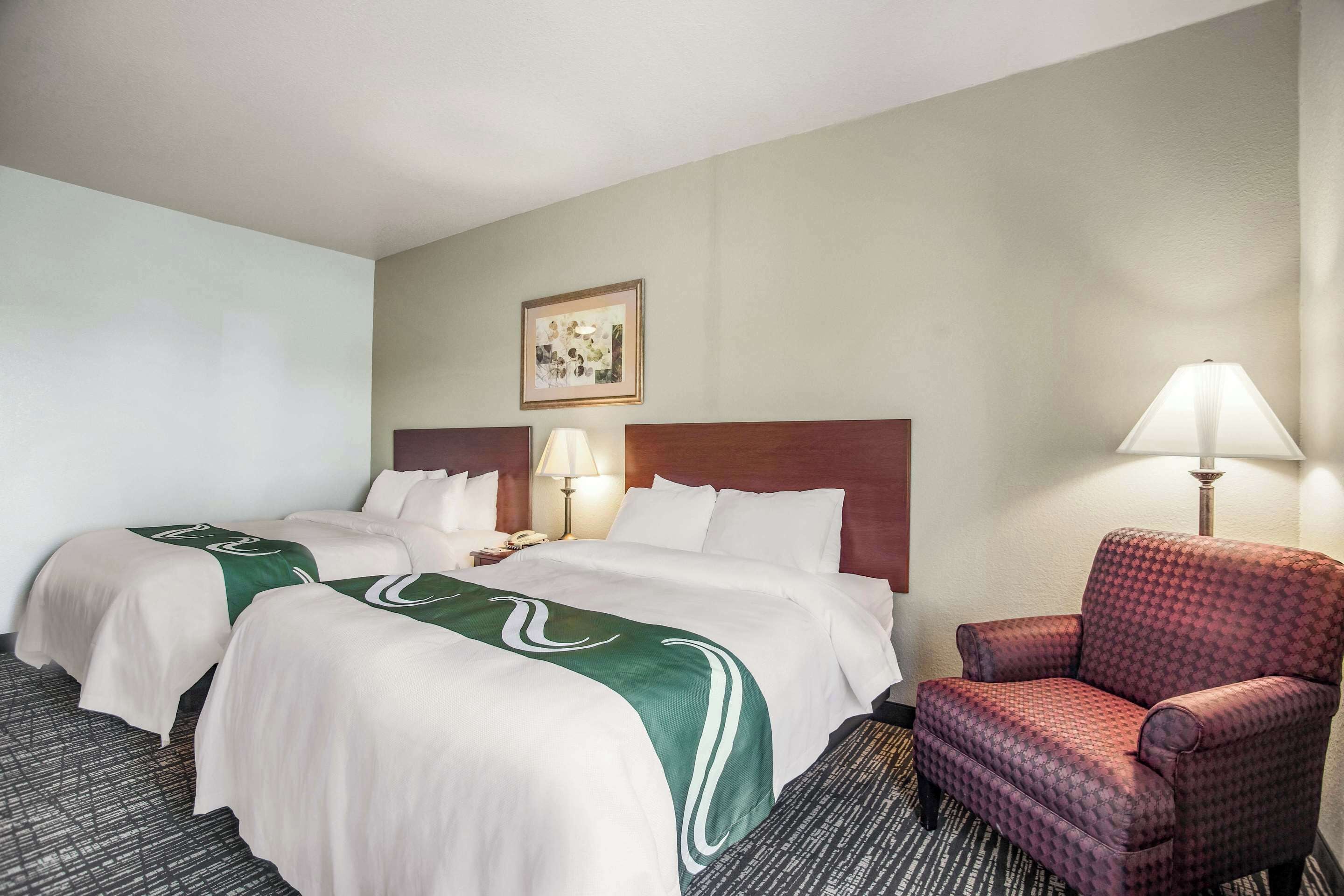A hotel room with two beds, a red armchair, a lamp, and a framed picture on the wall.