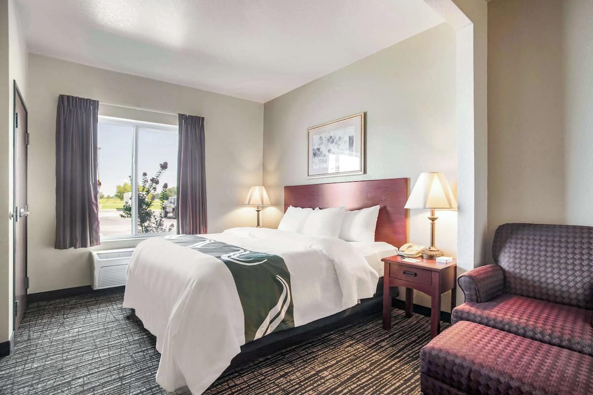 Une chambre d’hôtel avec un lit, une chaise, une table de chevet avec une lampe et une fenêtre avec des rideaux.