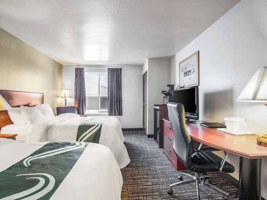 A hotel room with two beds, a desk with a lamp, a TV, and a window with curtains.
