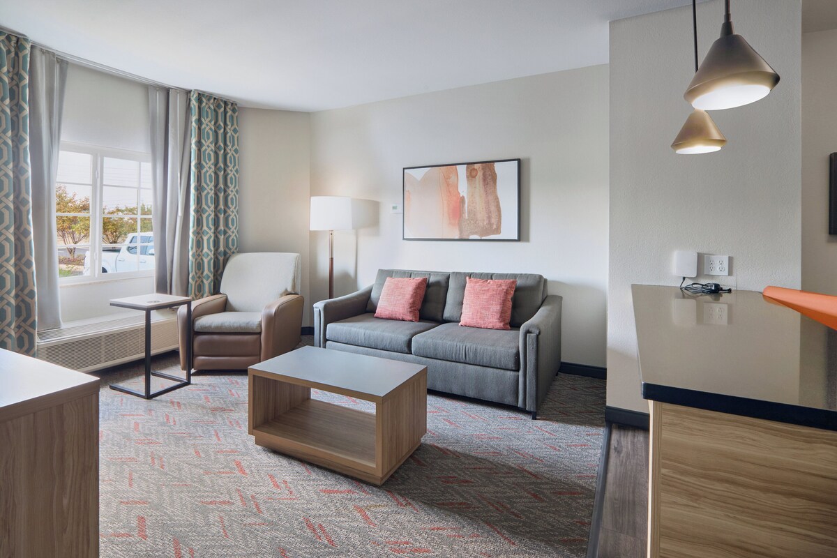 Ein modernes Hotelzimmer mit grauem Sofa, einem Holztisch, einem weißen Sessel und einem Schreibtisch mit Lampe.