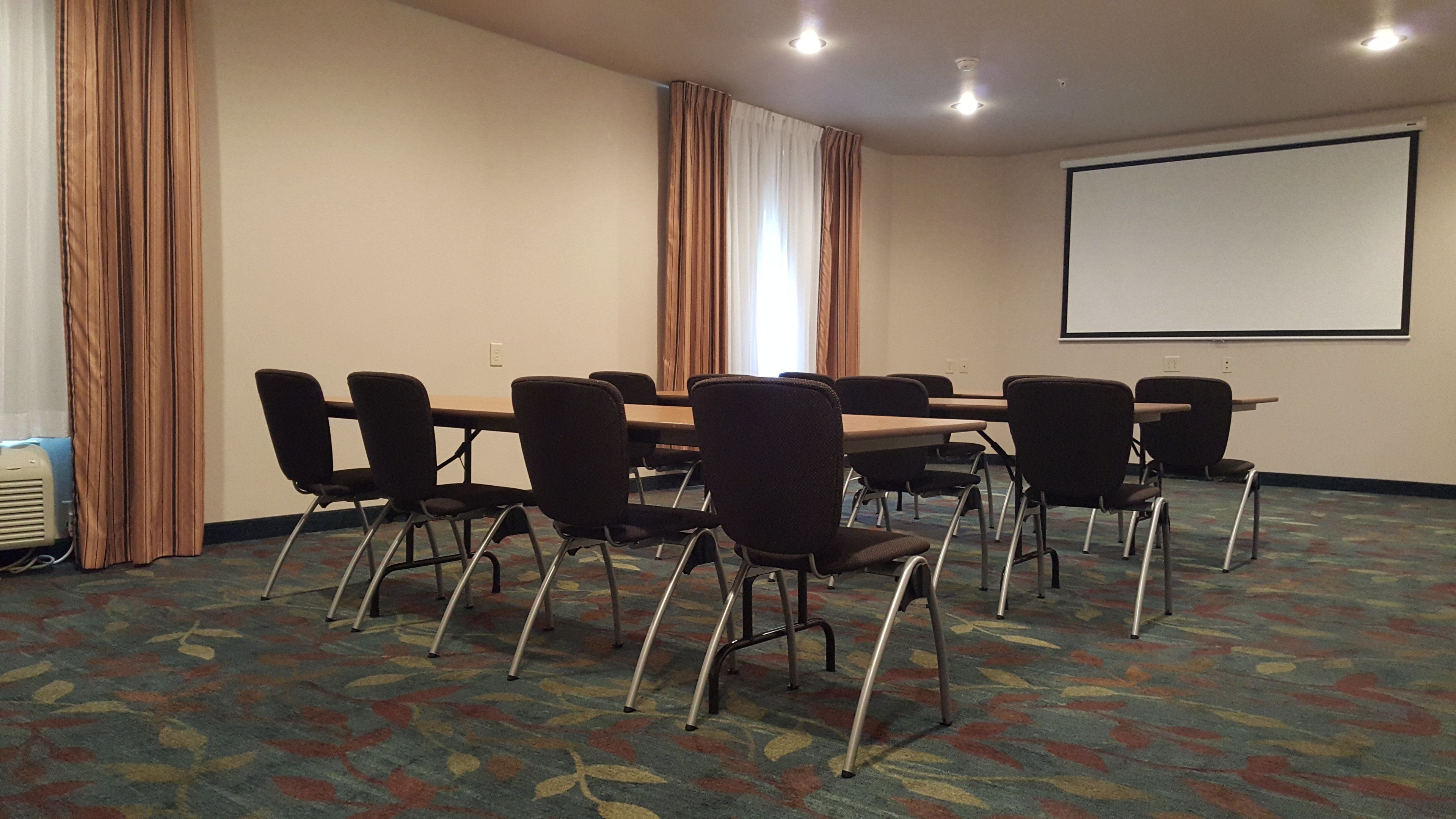 Un salón de conferencias con una mesa grande, sillas, una pantalla de proyector y cortinas.