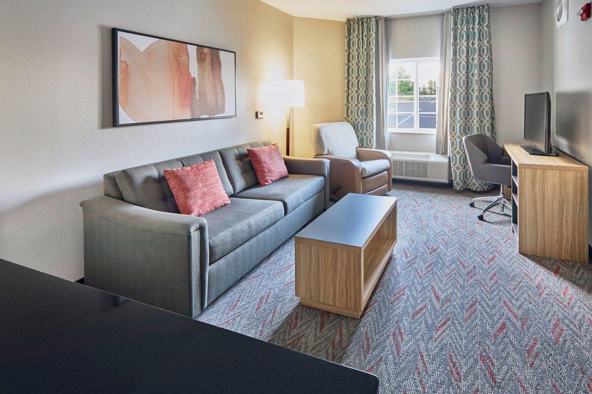 Une chambre d’hôtel avec un canapé gris, une table basse en bois, un téléviseur posé sur un meuble en bois et un tapis à motifs.