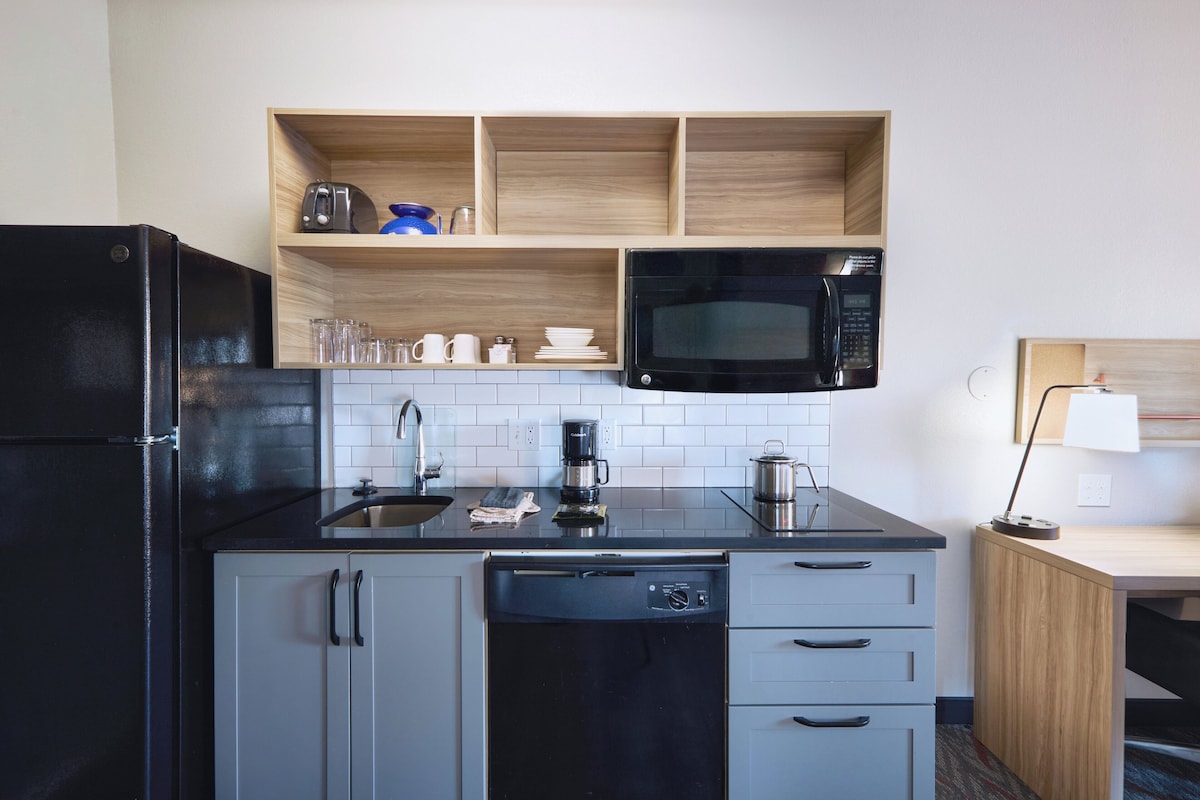 A compact kitchen with a black refrigerator, a microwave, a coffee maker, and a dishwasher. There are wooden shelves above the counter and a small cabinet below.