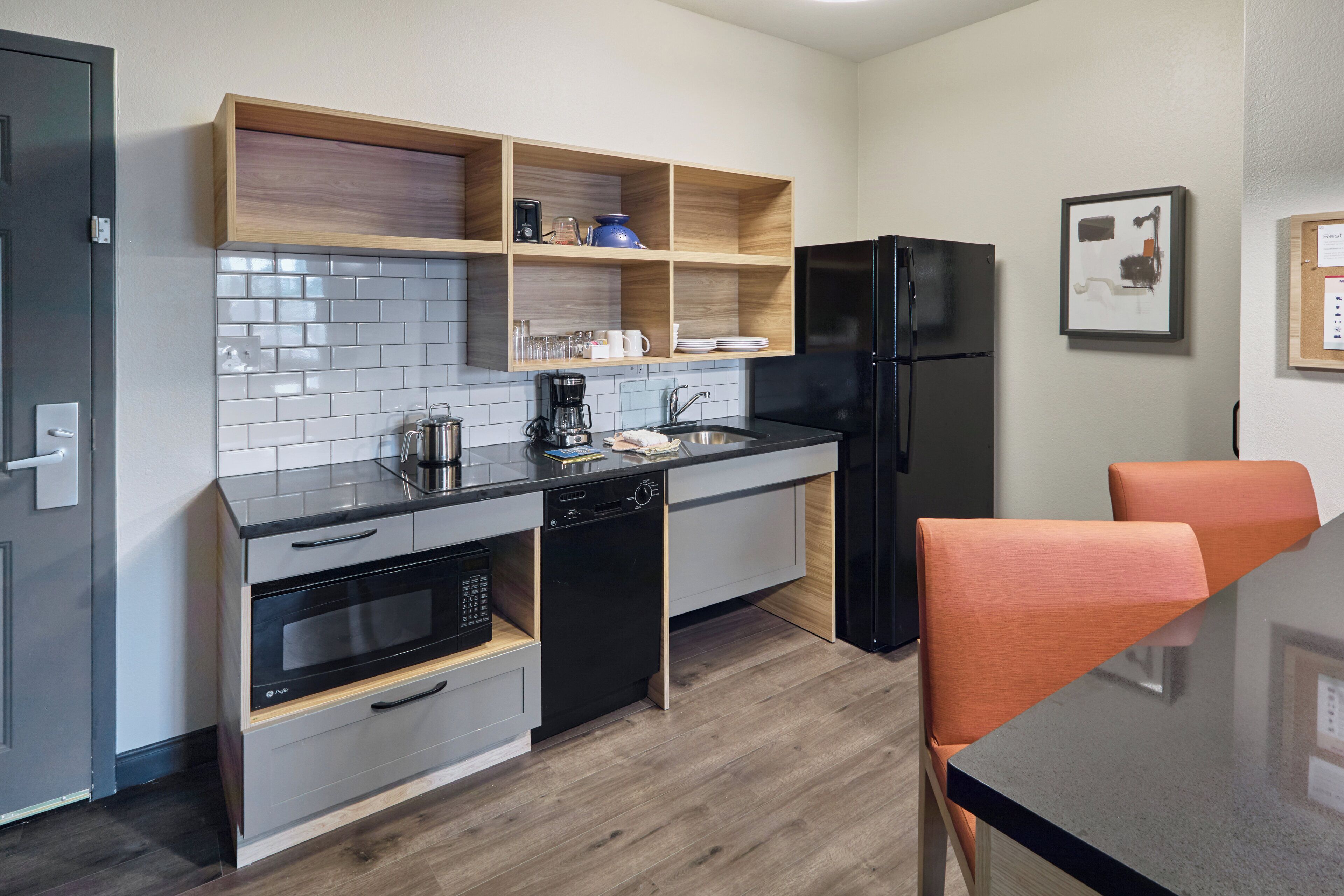 Una cocina compacta con lavavajillas, horno y refrigerador negro. Hay estantes de madera sobre la encimera y una mesa de comedor con sillas anaranjadas.
