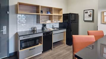 Una cocina compacta con lavavajillas, horno y refrigerador negro. Hay estantes de madera sobre la encimera y una mesa de comedor con sillas anaranjadas.