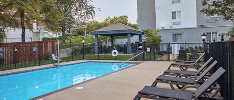 Una piscina con sillas de descanso, un pabellón y un edificio al fondo.